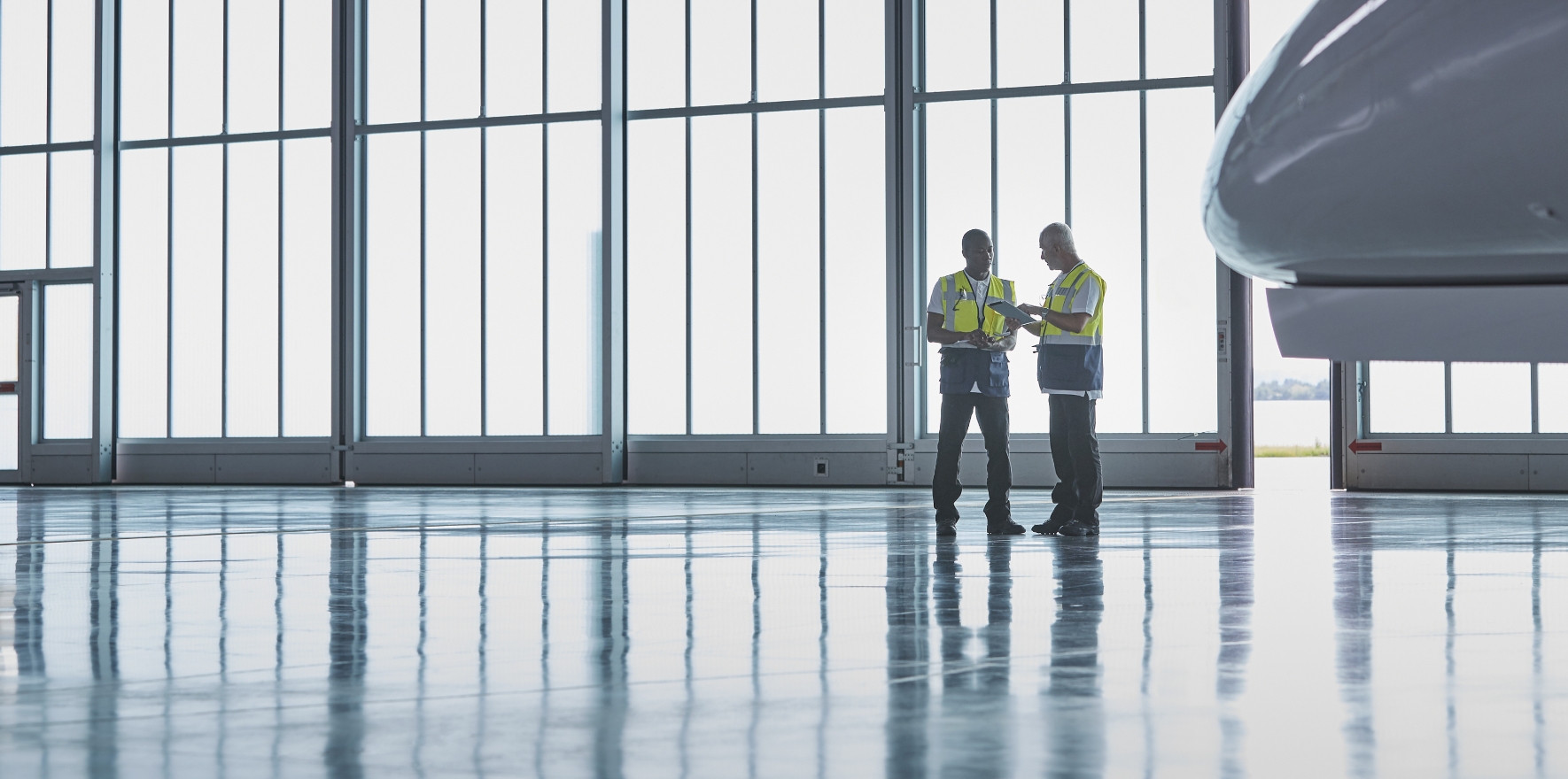 Two men wearing high visibility vests standing in an aircraft facility, reviewing papers on a clipboard
